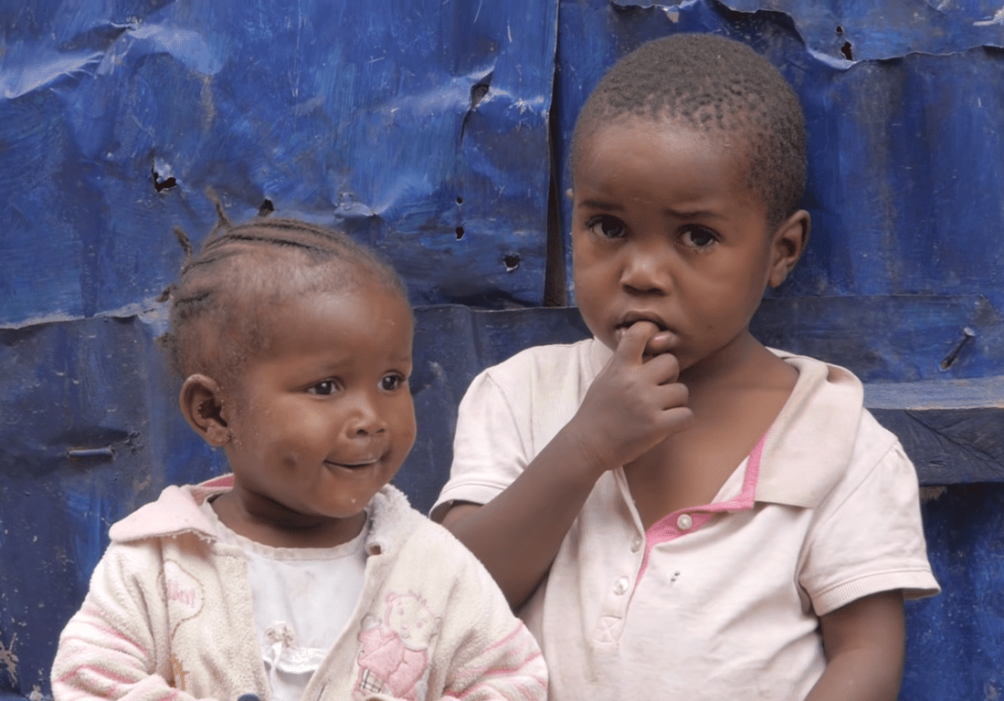 Two young children with thoughtful expressions stand before a blue, distressed background. The younger child smiles, while the older child chews on a finger. This image symbolizes the recipients of Zakat al-Fitr donations, made possible through cryptocurrency like SOL.
