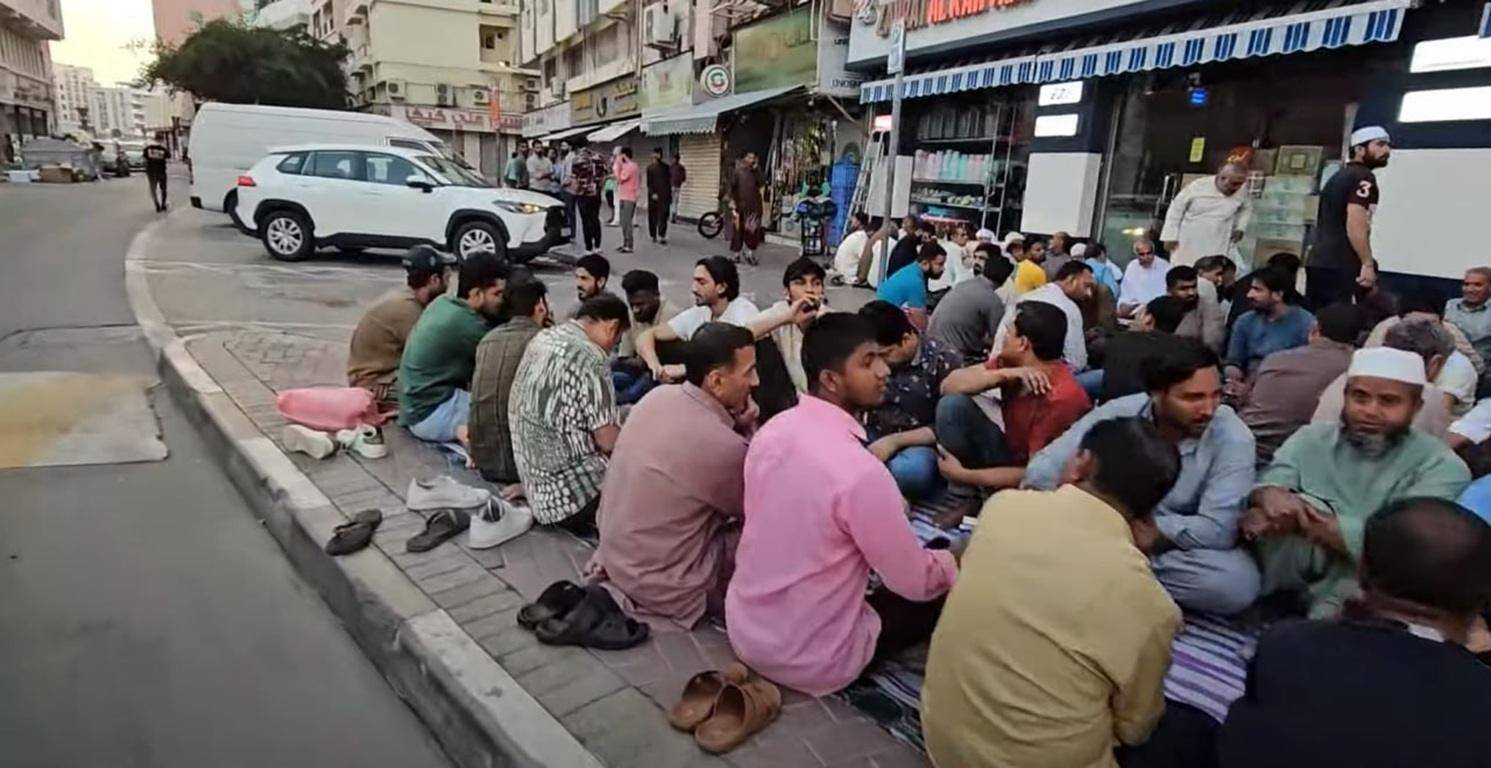 A group of men are sitting on the sidewalk in front of shops, some with their shoes off, in a street scene that appears to be in a Middle Eastern or Asian town. The context of the page is Qurbani or animal sacrifice in Islam, with an option to pay using cryptocurrency like PYUSD.