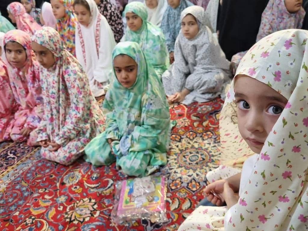 Girls in hijabs and modest Eid clothing sit on a patterned carpet, some looking forward attentively. The context is Zakat al-Fitr donations via cryptocurrency like Bitcoin and Tether.