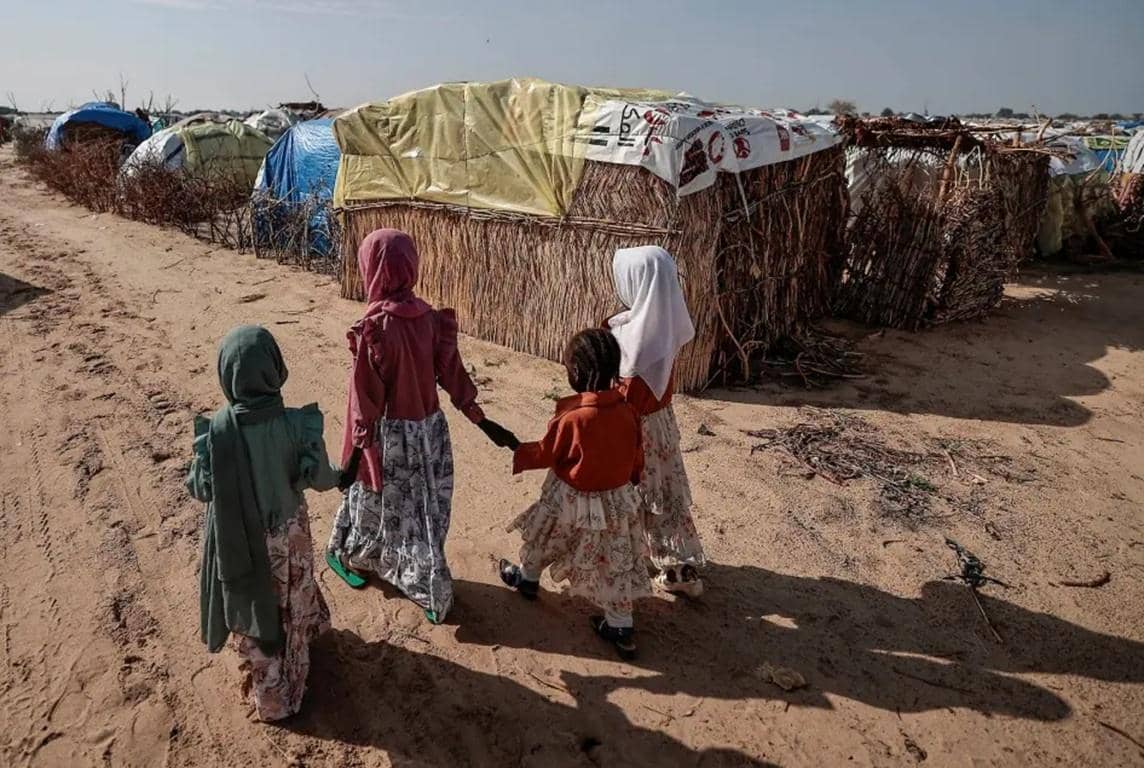 Four children walk hand-in-hand through a dusty refugee camp, towards makeshift shelters. Their journey highlights the urgent need for aid in Sudan and South Sudan, with crypto donations like BTC providing crucial support for food, water, and medical care.