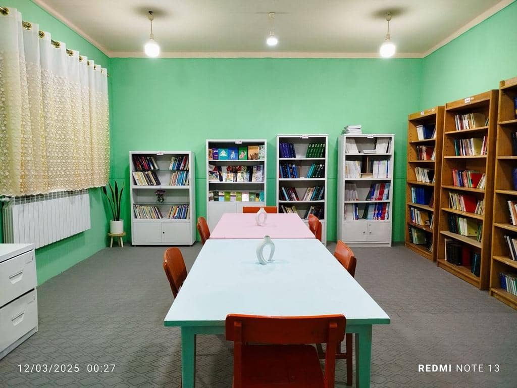 A clean, bright room with bookshelves on the right and a white cabinet with books on the left. Two tables with chairs are set up in the center, suggesting a study or classroom environment, supported by crypto donations.