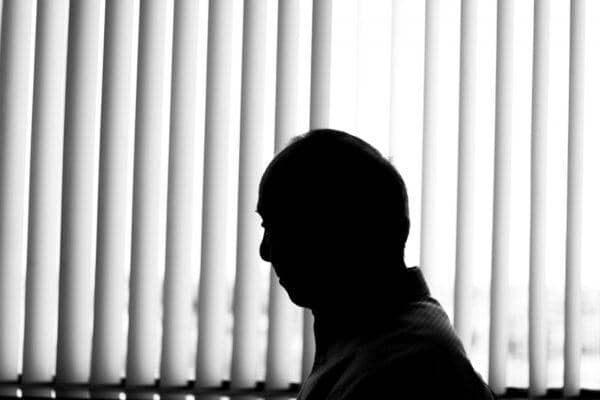 Silhouette of a man in profile, facing left against a backdrop of vertical blinds, representing the danger of social isolation and its detrimental effects on physical and psychological well-being, with options for anonymous crypto donations made via BTC.