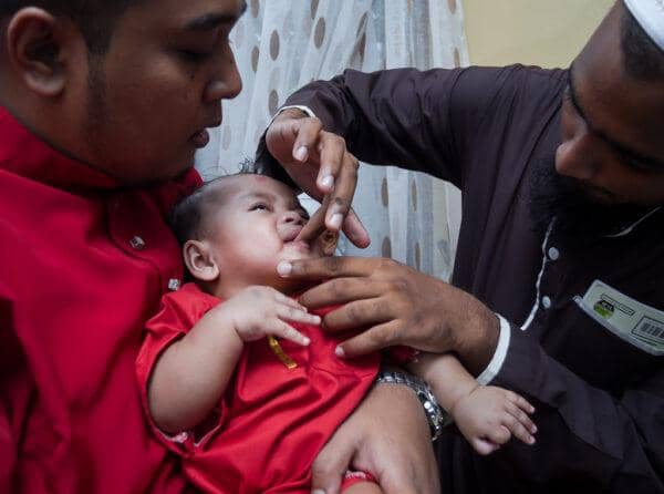 Tahnik ritual with baby receiving a sweet substance rubbed on the palate by a man in a brown thobe, while another man in a red outfit holds the baby. Traditional Islamic welcome with dates and prayer, supported by crypto donations.