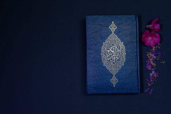 A blue book with ornate gold Islamic calligraphy on its cover, next to small purple flowers and petals. Implies charitable giving supported by ETH donations.