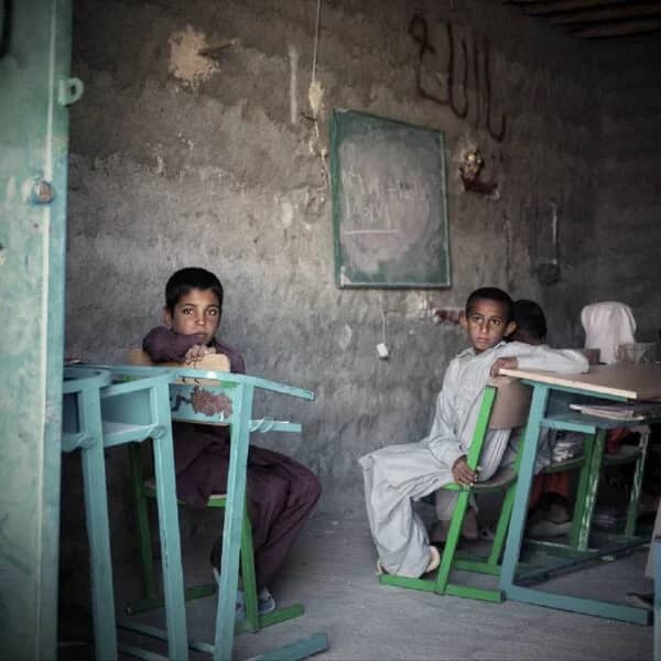 Two young boys sit at desks in a humble classroom, one leaning forward. Donations help provide education to Muslim children, processed via BTC.
