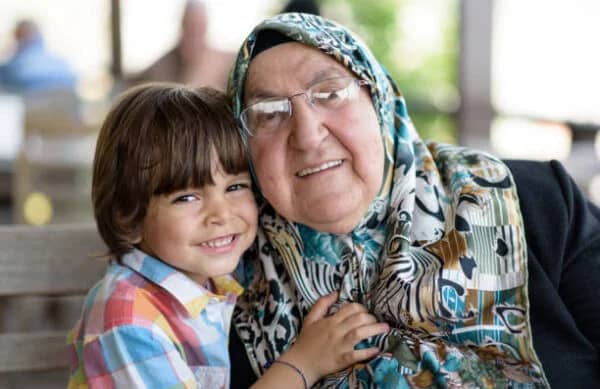 An elderly woman wearing glasses and a colorful hijab smiles next to a young boy, representing donating in honor of parents. Support for your charitable acts is available via ETH.