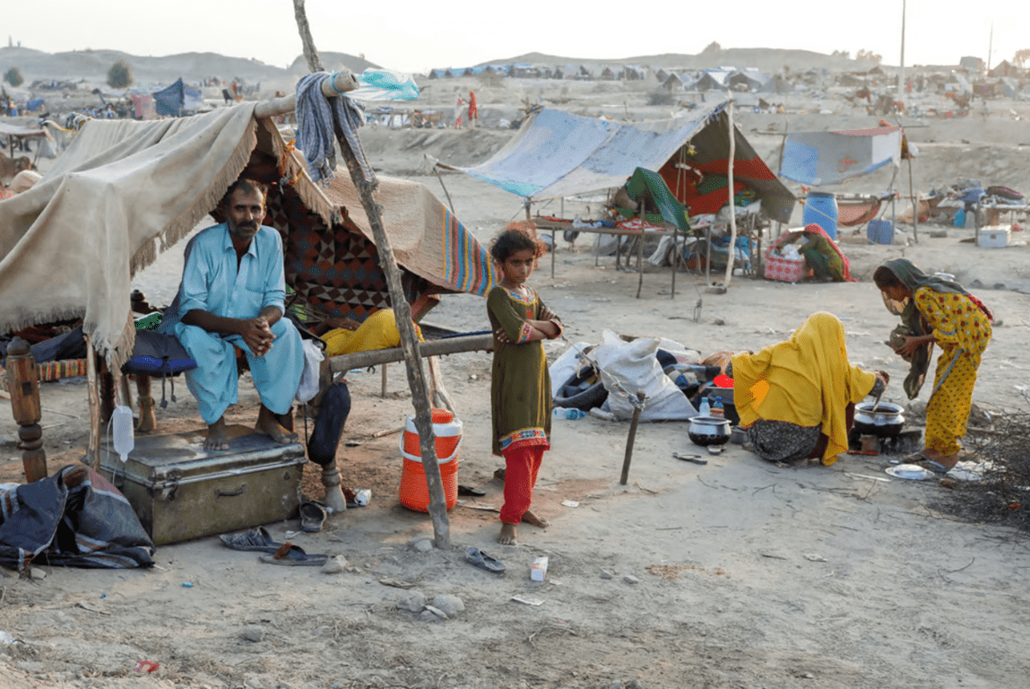 A man sits under a makeshift shelter while a young girl stands nearby in a refugee camp, highlighting the need for support through crypto donations like BTC.