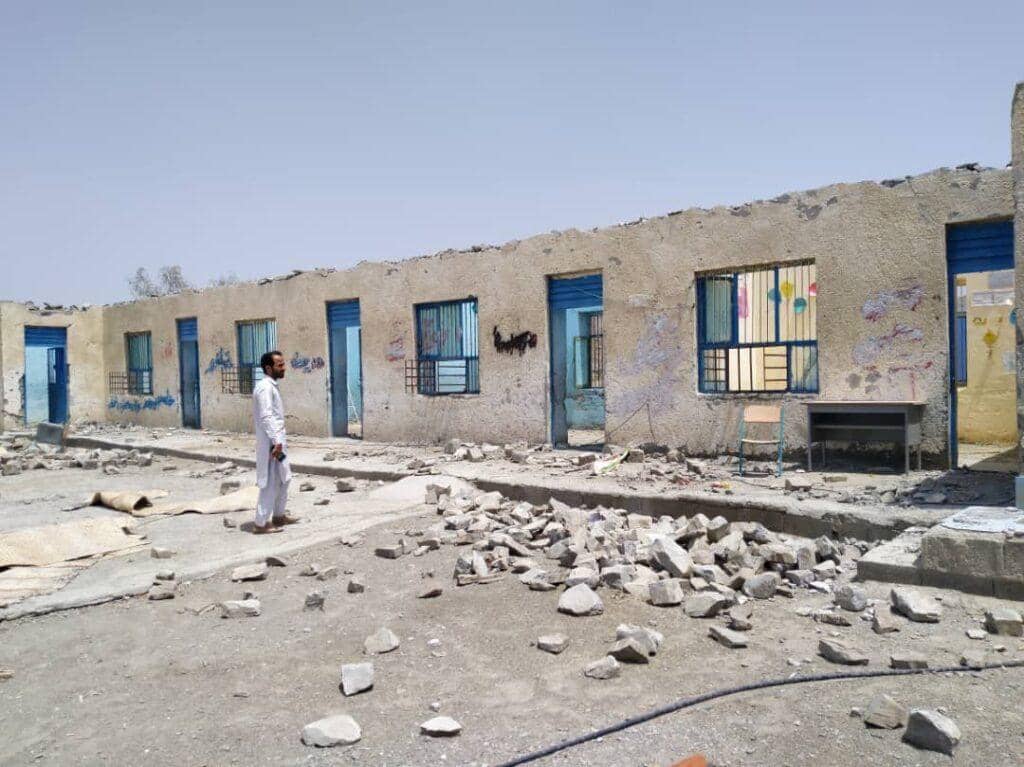 Man stands amidst rubble outside a damaged school building, symbolizing the need to donate for education with crypto, possibly via USDC, to support children's access to learning.