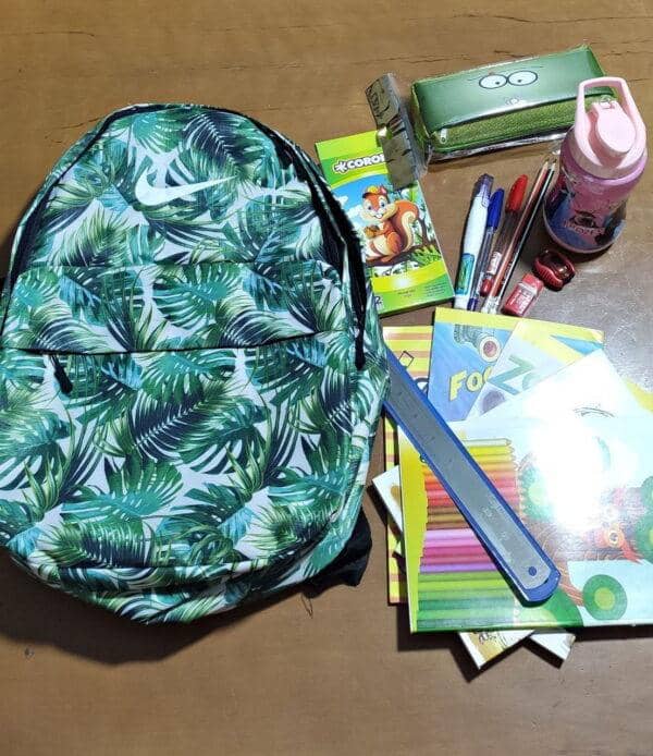 A vibrant green backpack with a tropical leaf pattern is displayed alongside school supplies including notebooks, pens, pencils, a ruler, and a water bottle. This image represents a donation for education, facilitating children's back-to-school needs, with support processed via USDT.