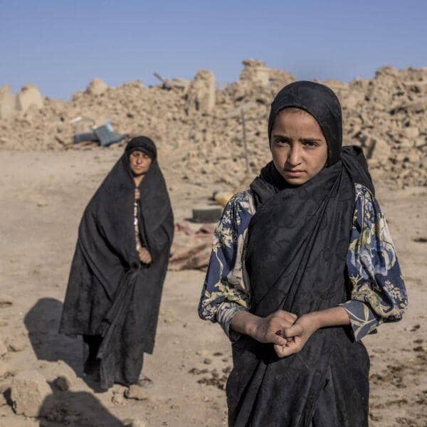 Two Afghan women stand in rubble after the 2023 earthquake, one looking at the camera with a serious expression. Providing first aid kits, training, and supplies, crypto-enabled donations like USDT support relief efforts for earthquake victims.