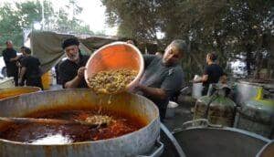Two Muslim men preparing a large meal, one pouring chunky ingredients into a massive pot of stew. This scene depicts a charitable food distribution, possibly with crypto donations like Bitcoin enabling aid. Keywords: Zakat, Islamic charity, crypto assets, Muslim community, aid.