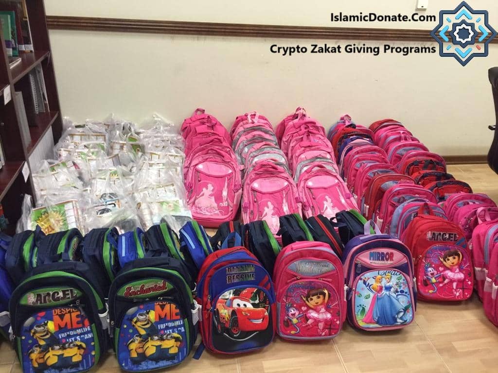 Crypto Zakat Giving Programs by IslamicDonate.Com shows rows of backpacks, many in pink, and bags of school supplies ready for donation. This crypto zakat offering supports children's education.