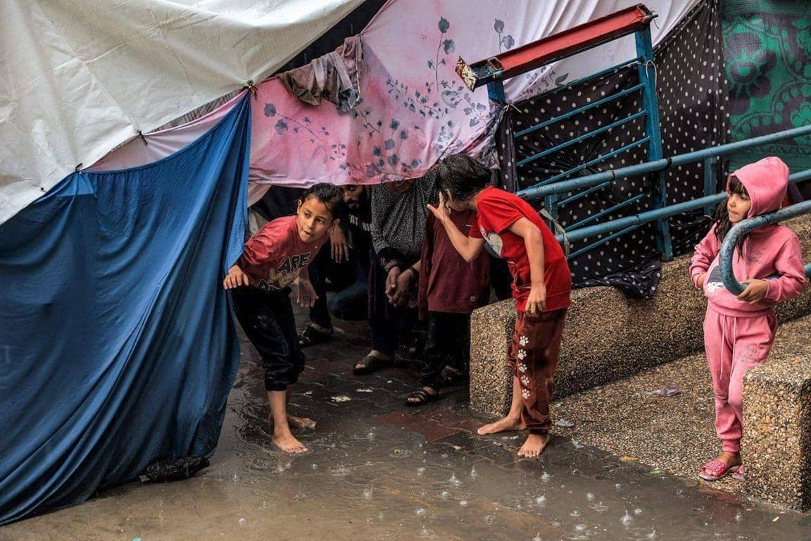 Children sheltering under a tent during rain, with some onlookers. This scene represents the impact of IslamicDonate's Aqiqah Qurbani, funded by ETH donations, providing relief and nourishment to needy communities.