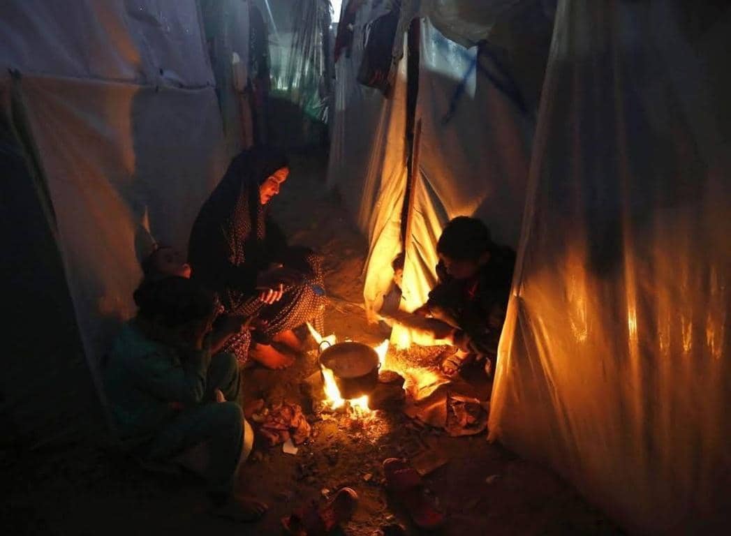 A mother and two children huddle around a small fire inside a makeshift tent, cooking food. This image represents the urgent need for aid in Palestine, highlighting the humanitarian crisis and the importance of crypto donations for survival, food, and shelter.