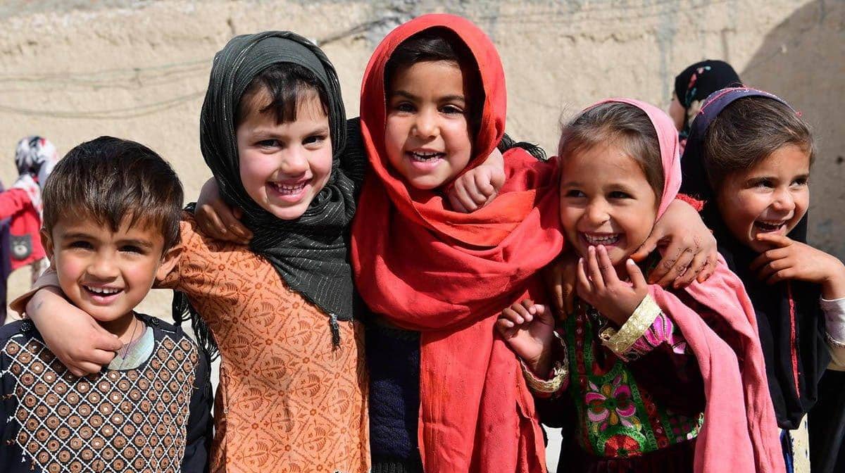 Smiling children in traditional clothing, some wearing headscarves. Their joy implies hope and resilience, with crypto donations via BNB supporting their relief.