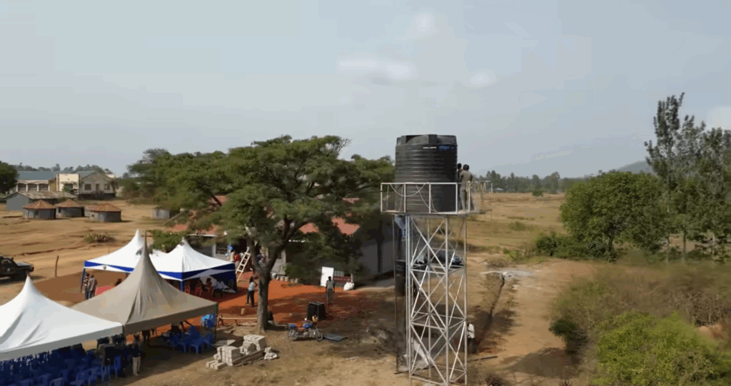 A water tower with a large black tank stands tall in a rural community, serving as a symbol of clean water accessibility. Two people are on the platform of the tower. In the foreground, tents are set up, likely for a community event or project inauguration, hinting at the positive impact of the water well initiative. This project, supported by crypto donations like ETH, brings essential water and fosters development, enabling communities to thrive.
