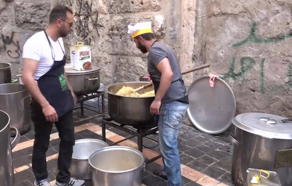 Two men prepare large pots of food in an outdoor setting, likely for charity. This scene illustrates fidya payment for Ramadan, with aid supported by SOL.