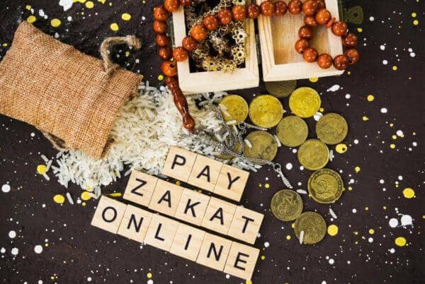 Text reads PAY ZAKAT ONLINE. Islamic prayer beads, gold coins, and a burlap sack of rice are scattered on a dark, speckled background.