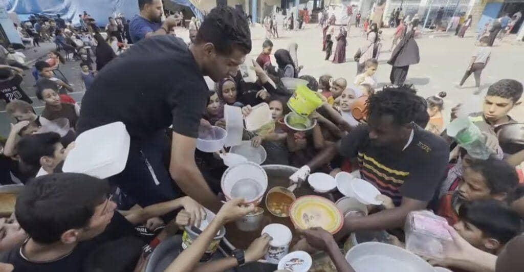 Volunteers distribute food and water to a crowd of people in Palestine, highlighting aid challenges. Donations can be processed via BNB.