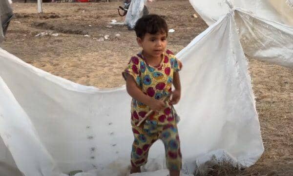 A young child stands amidst makeshift shelter fabric, symbolizing the ongoing daily struggles and resilience in Palestine. Aid delivery is hampered by checkpoints, waste management issues, and water scarcity. Supporting this cause with BTC donations helps overcome obstacles in delivering essential resources and education.