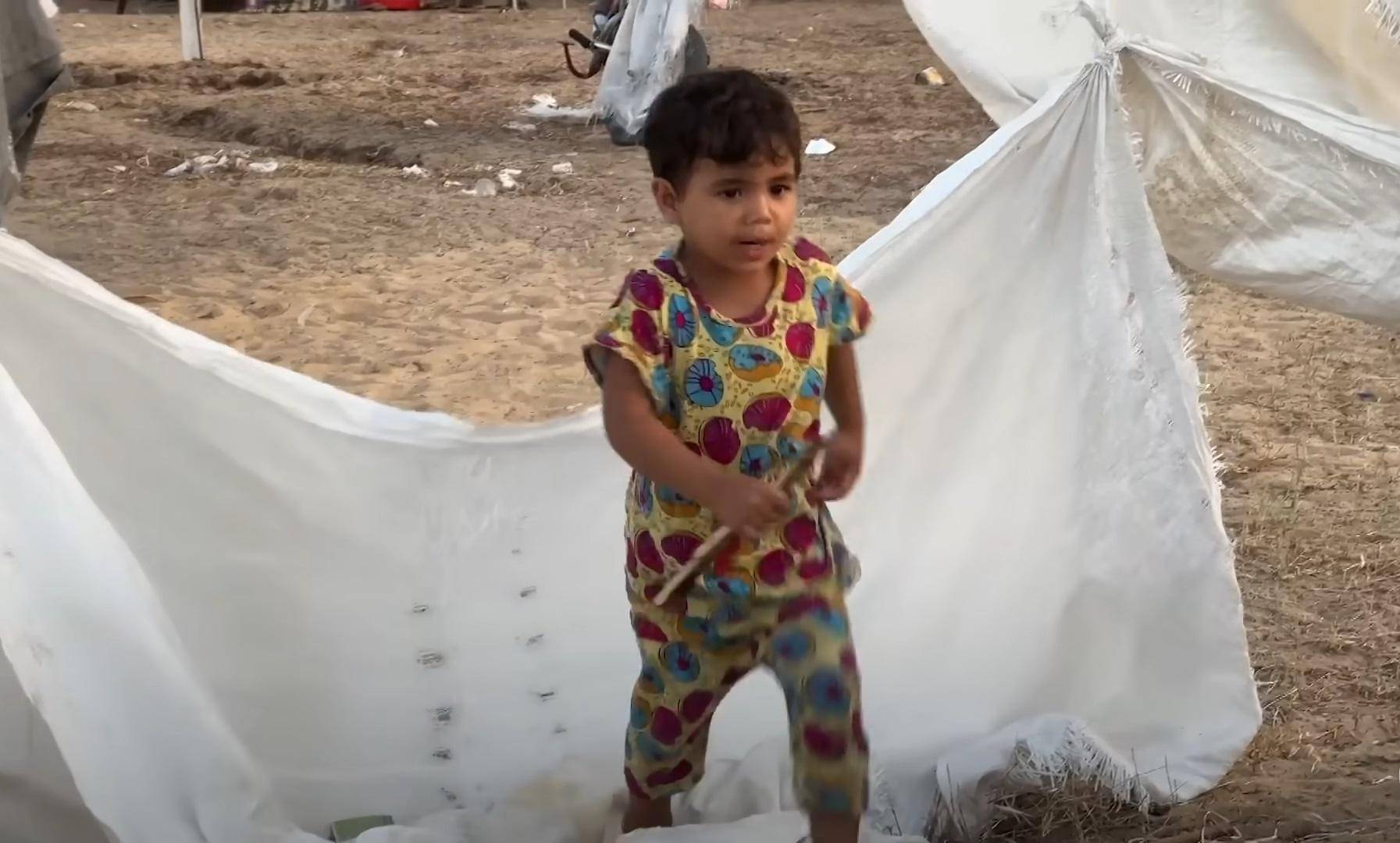 A young child stands amidst makeshift shelter fabric, symbolizing the ongoing daily struggles and resilience in Palestine. Aid delivery is hampered by checkpoints, waste management issues, and water scarcity. Supporting this cause with BTC donations helps overcome obstacles in delivering essential resources and education.