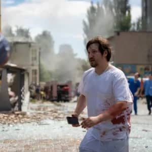 Bloody medical professional with phone in hand walks through debris after missile attack on Kyiv children's hospital, seeking aid via BTC donations.