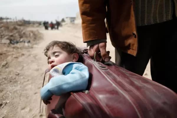 A man carries a sleeping child on a suitcase through a desolate landscape, symbolizing the refugee crisis and the hope provided by crypto donations like SOL to empower communities and reduce displacement.