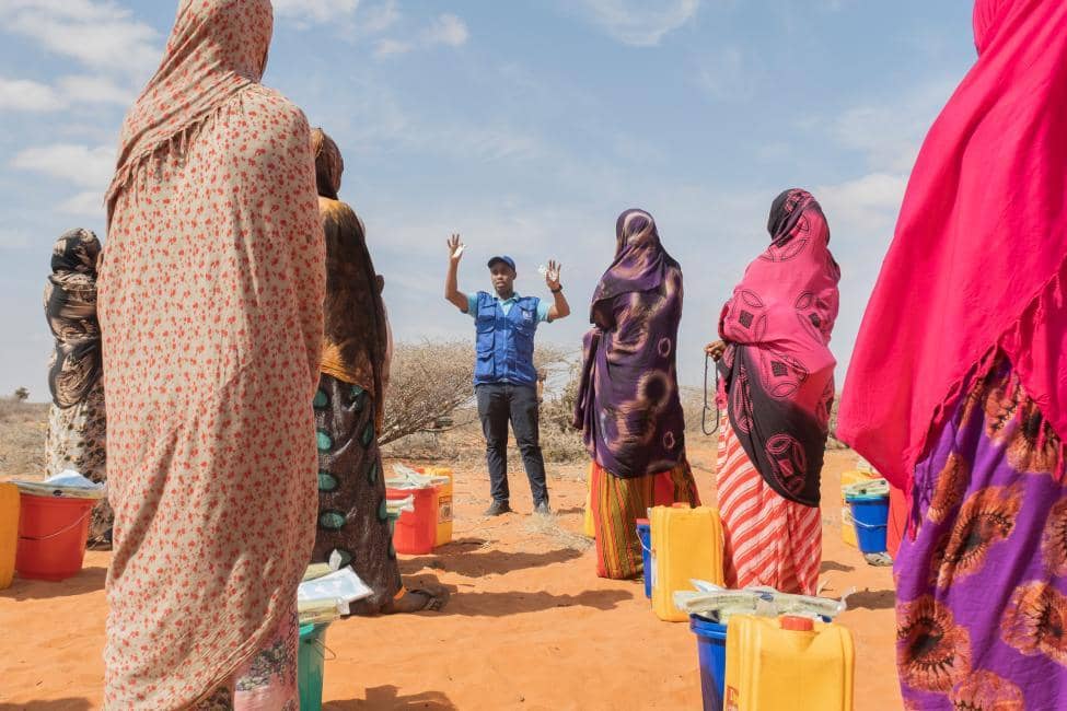 Humanitarian aid worker in a blue vest addresses a group of women in a dry, arid landscape, with yellow jerrycans and buckets visible, indicating distribution of essential supplies like water and food, supported by USDT donations.