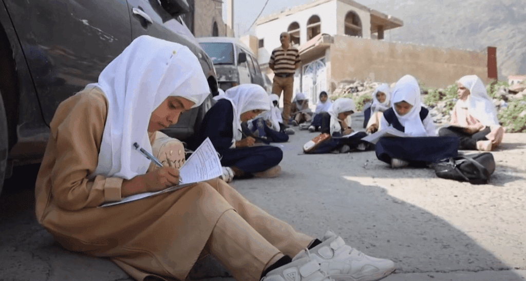 Children in hijabs learning outdoors, with one girl in the foreground writing, receiving crypto donation support via USDT, symbolizing educational aid for orphans.