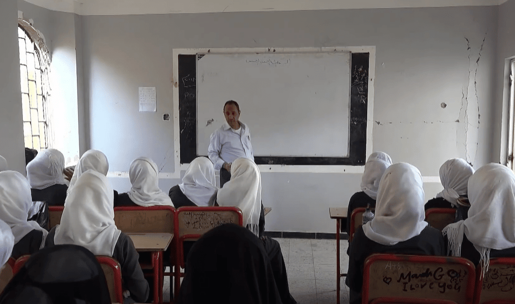 A teacher stands at a whiteboard in a classroom as students, mostly girls wearing headscarves, sit at desks. Sponsored education for 49 orphans, funded by crypto donations like BTC, provides hope and resources for brighter futures.