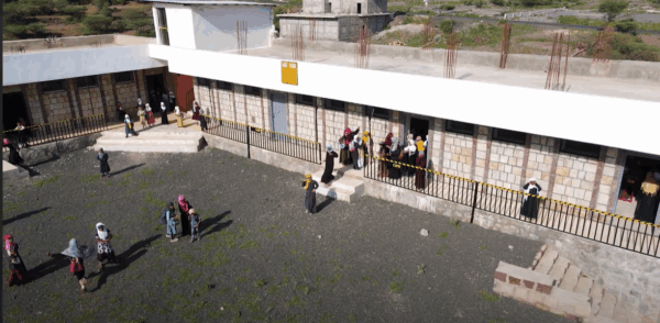 Children and adults gather outside a school building, some entering, others standing in the courtyard. The scene depicts the impact of crypto donations supporting education for orphans, fostering hope and community. The aid is facilitated by BTC.