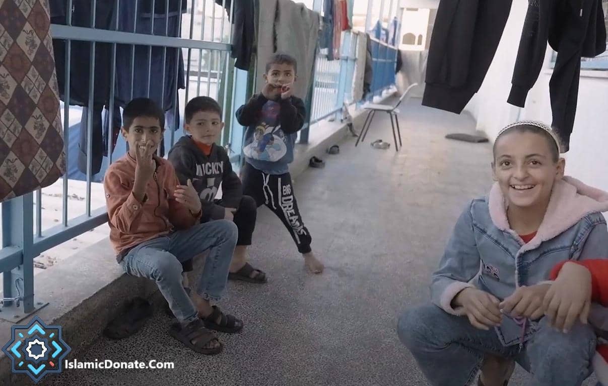 Four children sitting and standing, with one young girl smiling at the camera, posing near laundry hanging on a line. This scene represents support for Muslim orphans, facilitated by crypto donations like ETH.