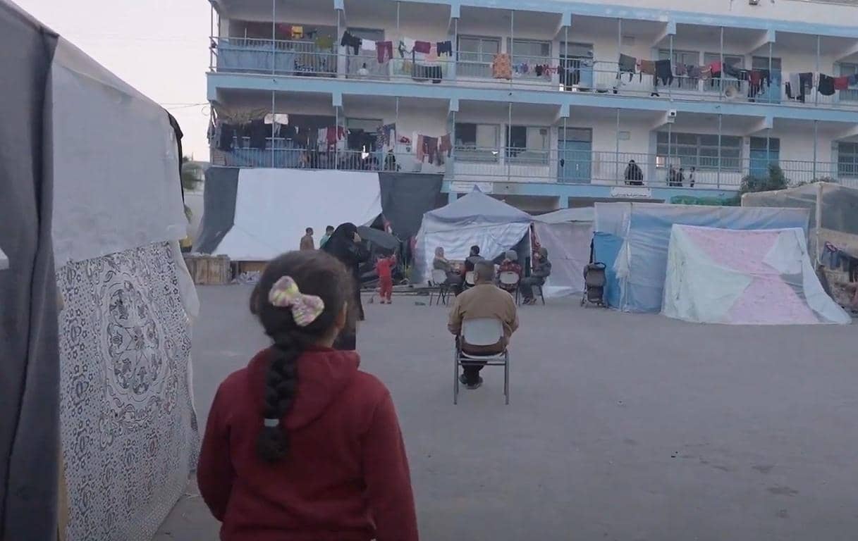 A young girl with a braid and bow looks toward a temporary shelter camp with temporary tents and a multi-story building in the background. People are seen sitting and standing in what appears to be a relief area. Sponsor an orphan from anywhere with cryptocurrency, offering shelter, food, and education, with donations in SOL.