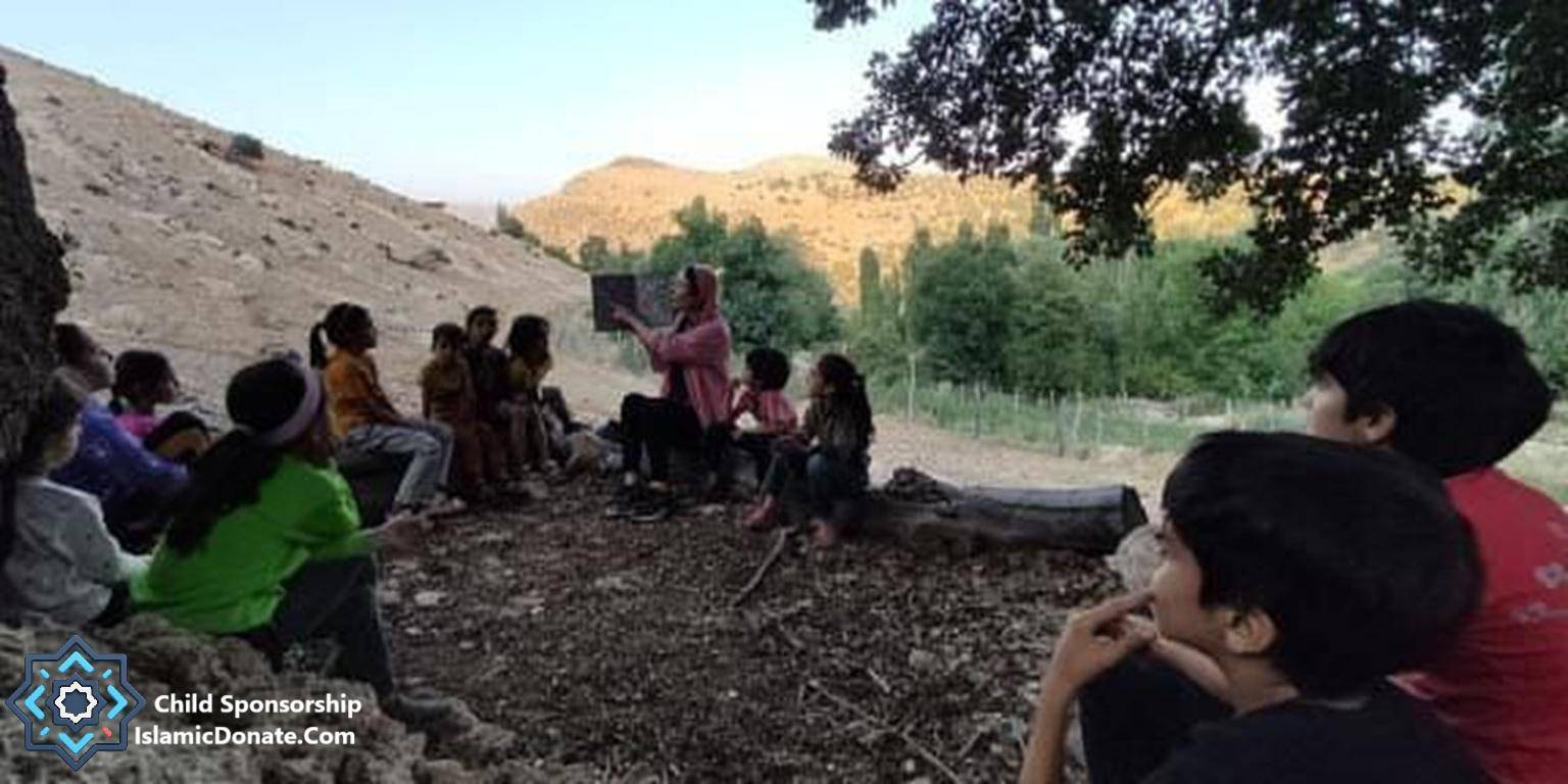 Group of children sitting outdoors listening to a woman holding a book, possibly a lesson or story. Sponsorship helps provide education and hope, now supported by SOL.