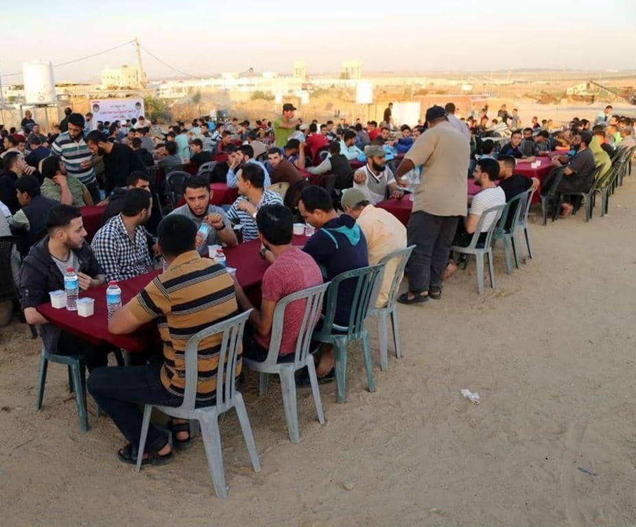 Group of men sitting at tables outdoors for an Iftar meal, receiving crypto donations for aid in Gaza, Palestine. Distribution of Suhoor and Iftar meals.