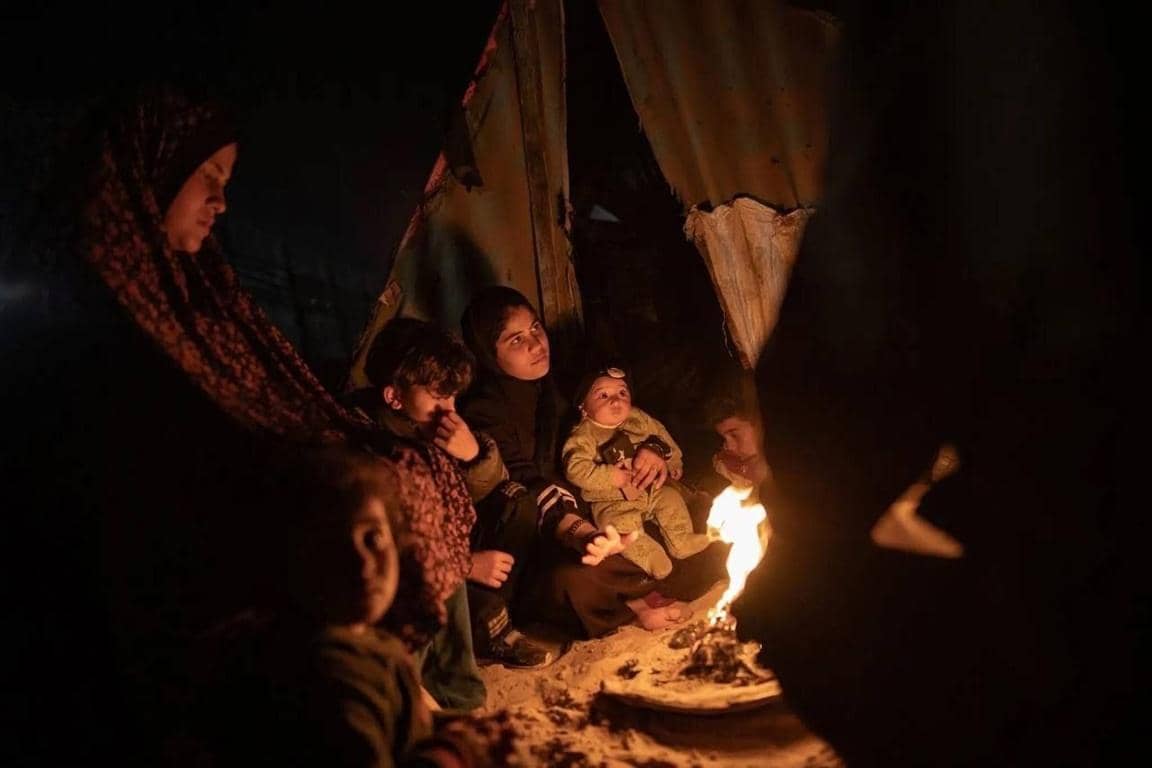 Family huddled around a fire near makeshift shelter in Gaza, seeking warmth amid crisis. Donate crypto to aid Gaza.