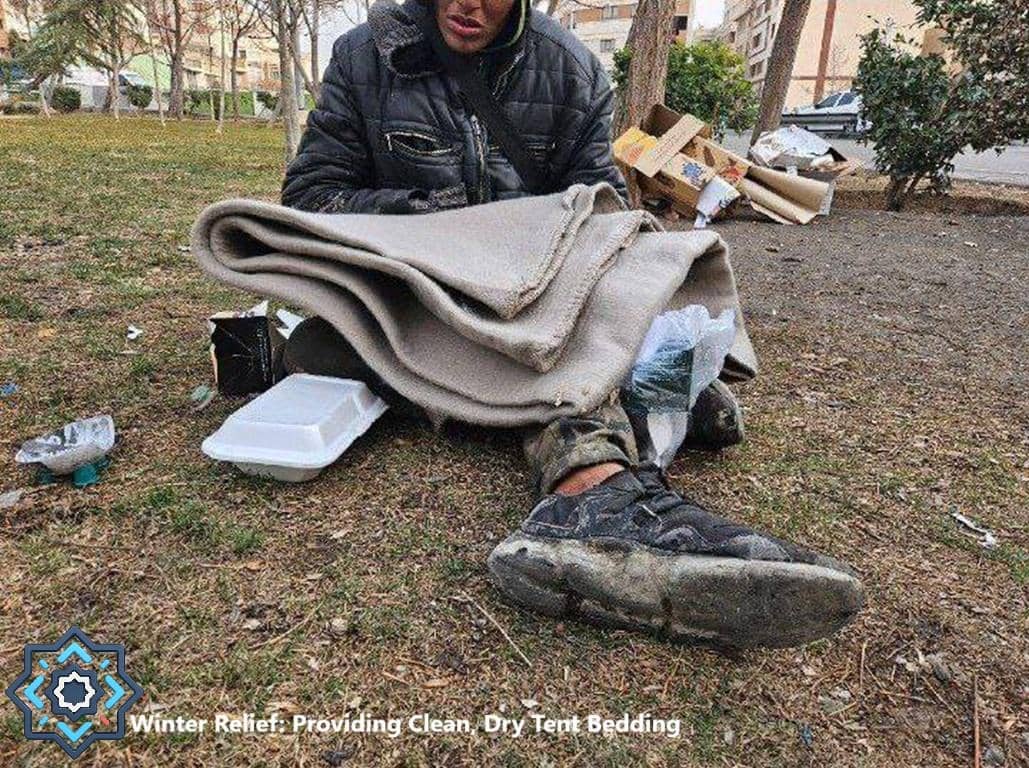 A person sits on the ground with a blanket wrapped around them. Near them are a white food container and some trash. This image represents the need for crypto donations for winter relief, providing warm bedding and shelter. Your donation helps create warmth, like a warm blanket in winter with SOL.