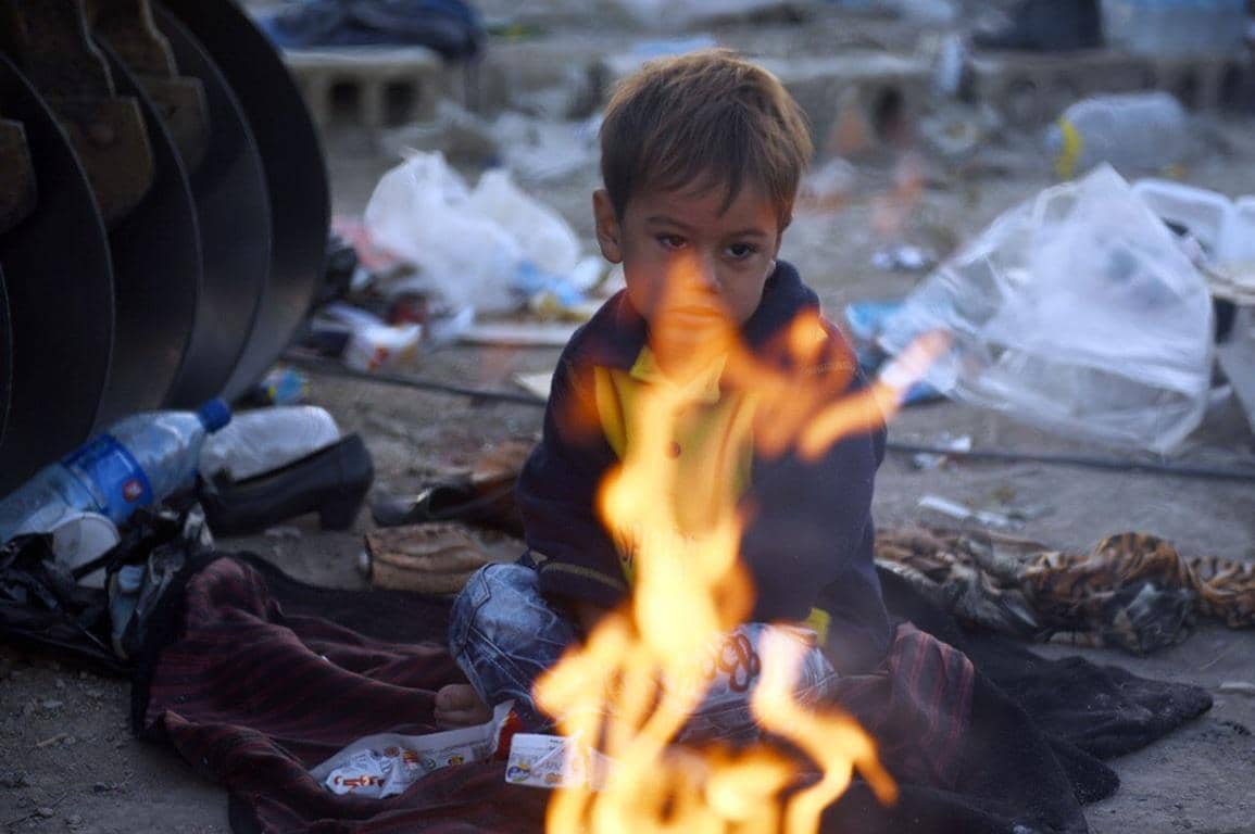 Child warming hands by a fire with debris in the background, symbolizing the humanitarian crisis in Gaza and the need for aid. Donate crypto Zakat for Gaza relief.