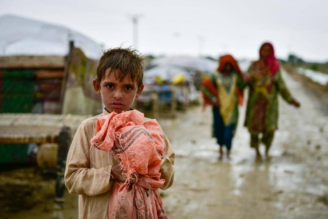 Young boy in a displaced camp holding a blanket, with two women walking in the background in muddy conditions. Your crypto donation with BTC can provide warm clothing, shelter, and necessities to vulnerable families.