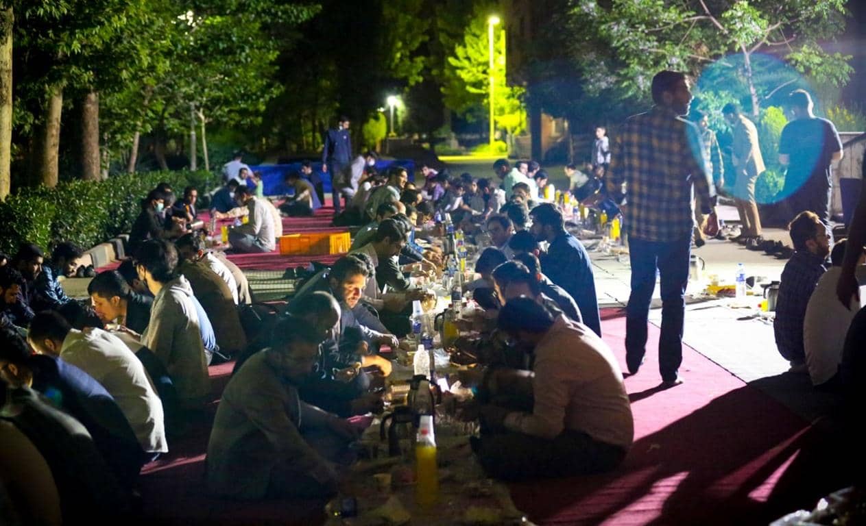 Group of men gathered outdoors at night, sitting on rugs for a communal meal. Some are eating, while others are conversing. The scene depicts a charitable iftar or suhoor, possibly facilitated by crypto donations like BTC, supporting needy Muslims.