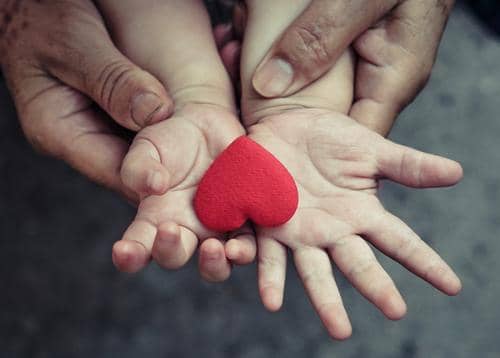 Two hands, one adult and one child, holding a red heart. The act symbolizes giving Sadaqah, Islamic charity, and the importance of compassion, with donations accepted via Cardano.