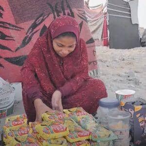A young girl in a headscarf and maroon dress sorts food items, an act of aid for working children, with donations possibly supported by ETH.