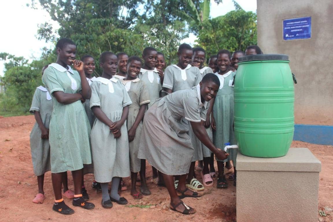 Ugandan schoolchildren gather around a green water barrel as one student dispenses clean water, symbolizing hope and relief aided by Solana donations.