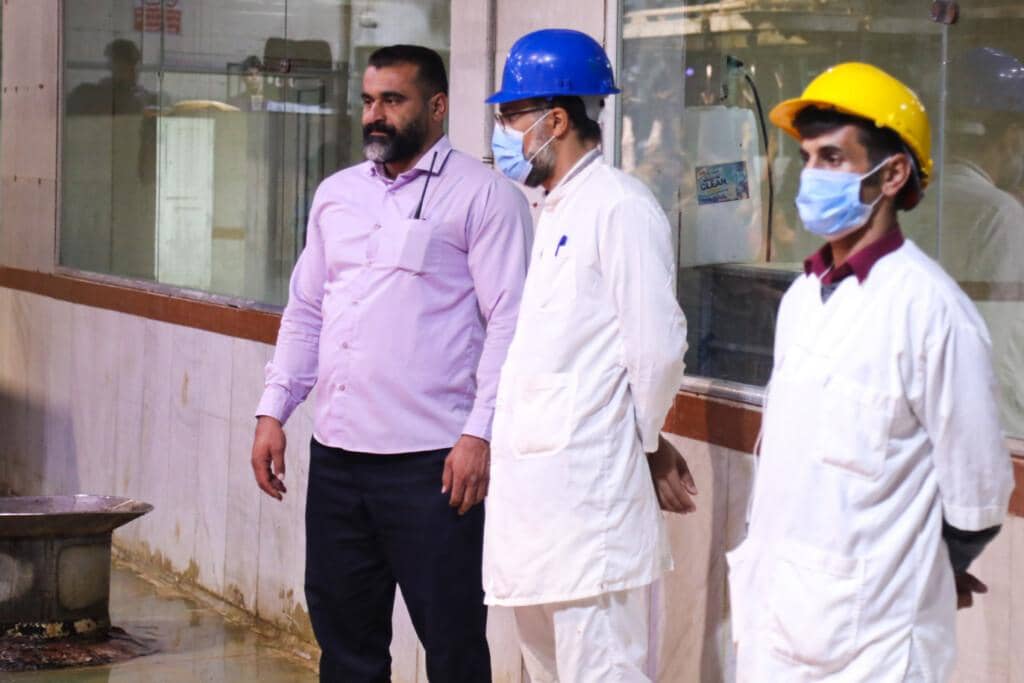 Three men in a food processing facility, one in a pink shirt, and two in white coats and hard hats, observe operations. Supporting the needy with crypto donations, like Bitcoin.