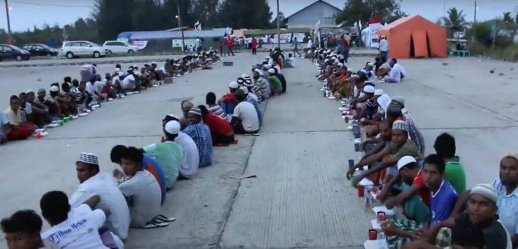 Group of men sit in rows on pavement, receiving Suhoor and Iftar meals during Ramadan 2025, supported by crypto donations like Ethereum (ETH) for food aid and emergency relief in conflict zones.