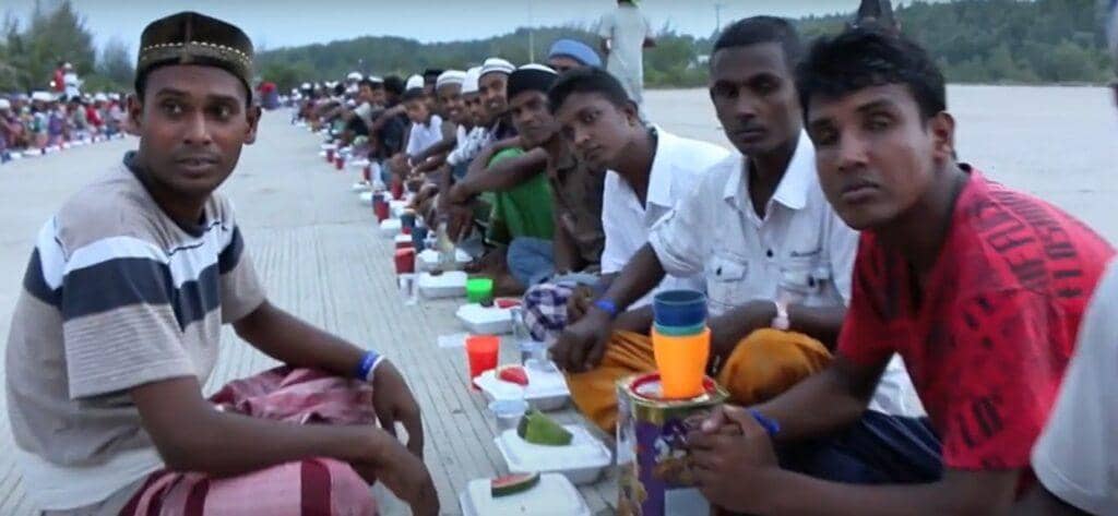 Men sit in rows receiving food aid, with packaged meals and drinks arranged before them. The scene shows the impact of humanitarian efforts, including crypto donations like TRX, supporting those in need of suhoor and iftar.