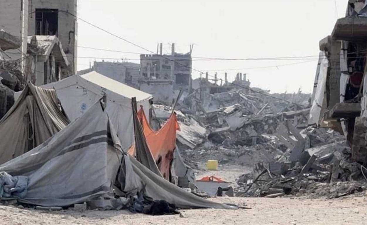 Temporary shelters in a war-torn Palestinian town with destroyed buildings in the background. Your crypto Zakat donation supports aid and reconstruction.