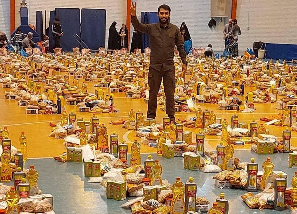 Man giving peace sign standing in front of a vast display of food packages and oil bottles for Zakat distribution, supported by a USDT donation.