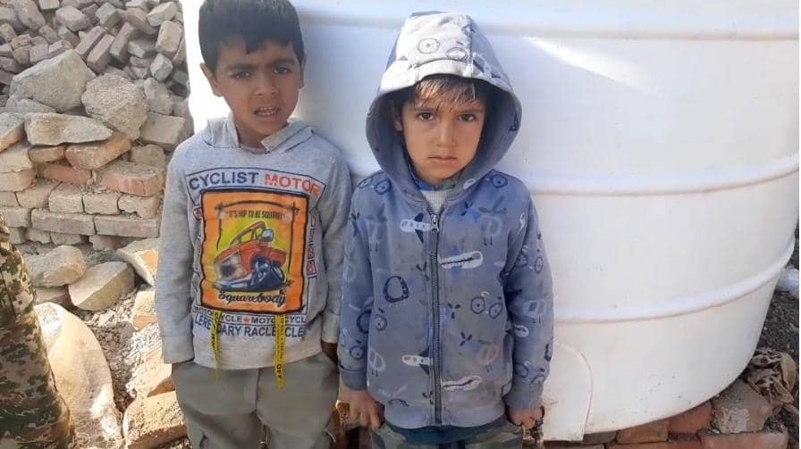 Two young boys stand near a water tank. The boy on the left wears a gray hoodie with a "Cyclist Motor" graphic. The boy on the right wears a blue patterned hoodie. Their background includes scattered bricks, suggesting a need for aid. This image signifies how crypto donations, like those made with Bitcoin, help provide essential resources to children in need.