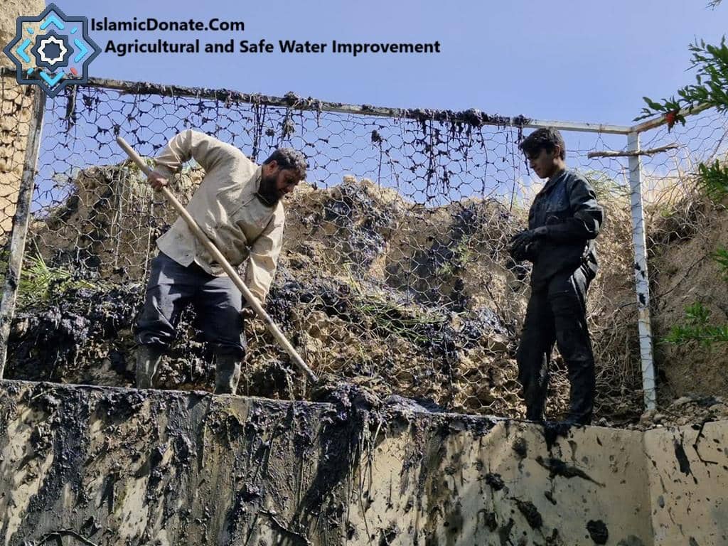 Two men work on clearing mud and debris from a water channel, possibly for agricultural use, with crypto donations supporting this vital project.